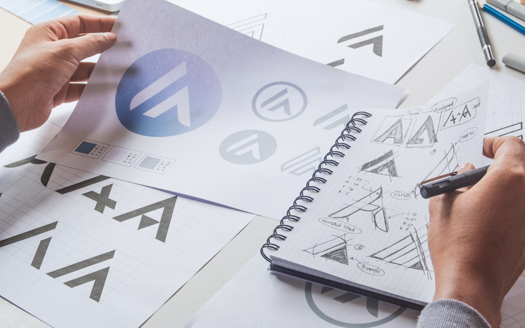Two hands on top of a sketchbook and several loose sheets of paper, designing variations of a logo.
